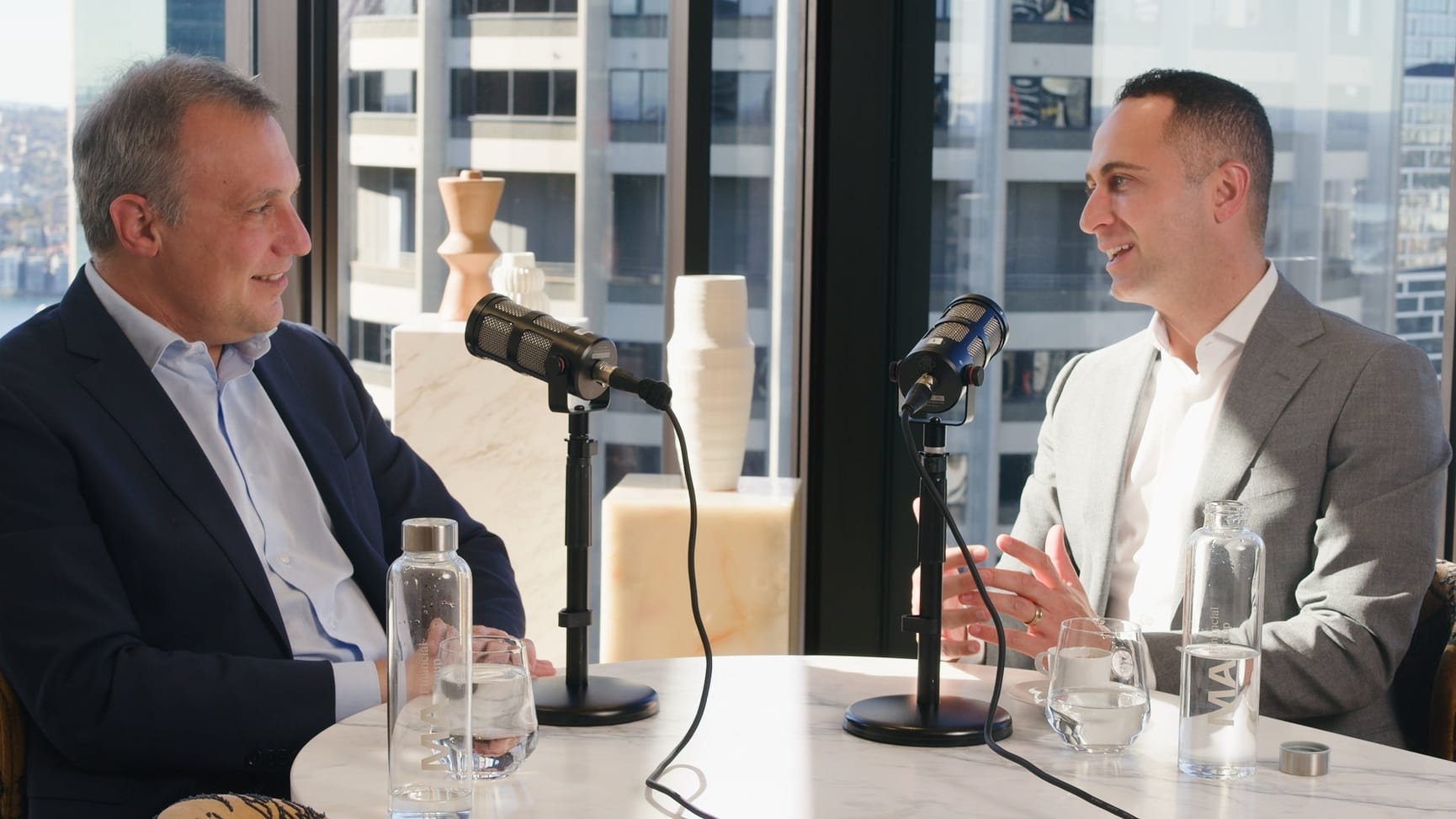 Two men sitting in front of microphones with a window showing Sydney skyline in the background