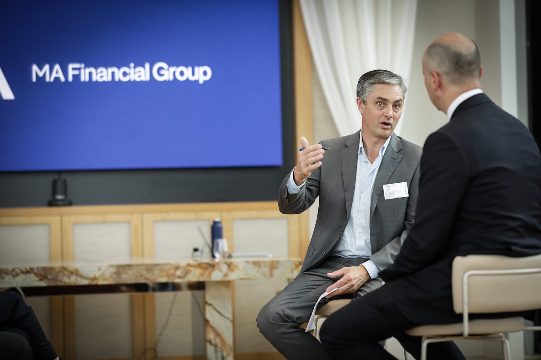 Two men in discussion with MA Financial Group logo in background on a plasma screen