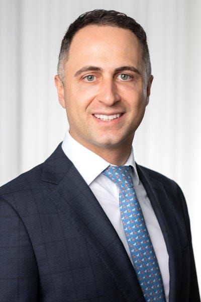 Frank Danieli wearing a check charcoal suit with a blue tie and white business shirt in front of a white draped curtain.